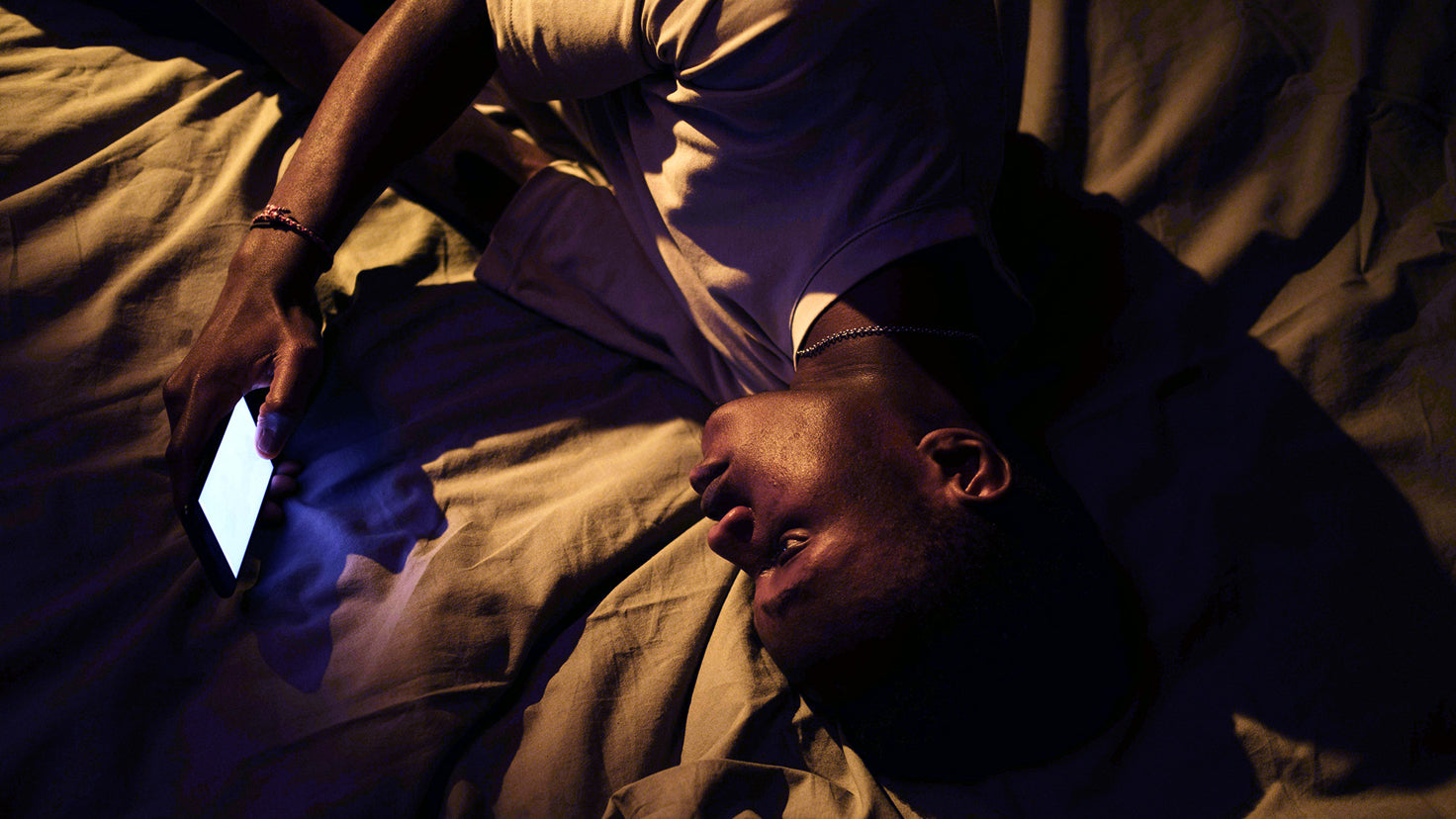 A young man lies in bed awake, the screen from his phone illuminating his face.