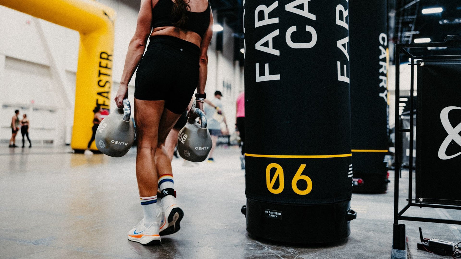 A woman walks away from the camera carrying two sixteen kilogram kettlebells, ready for the farmers