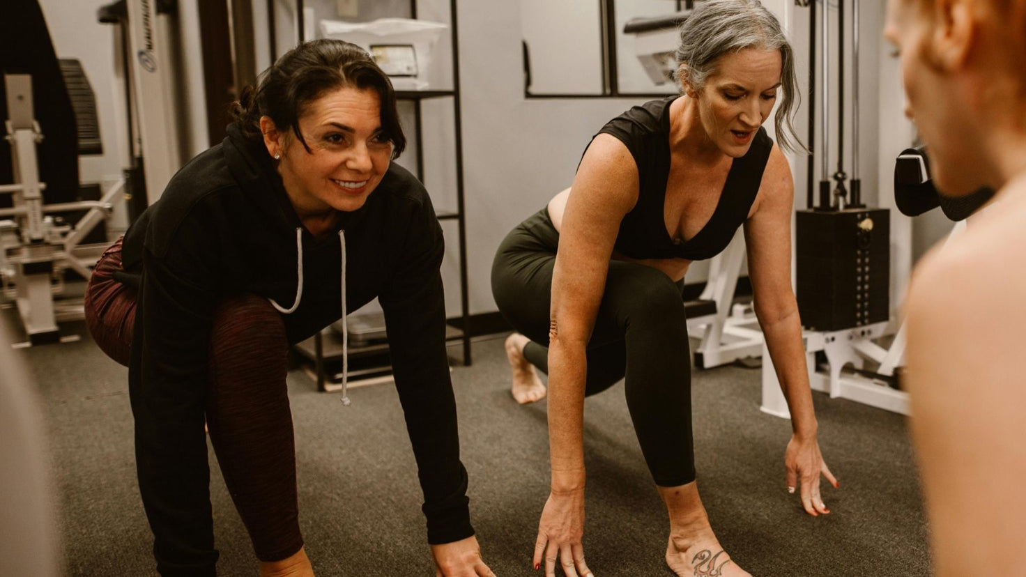 Two women are coached in a small gym, and performing deep forward lunges.