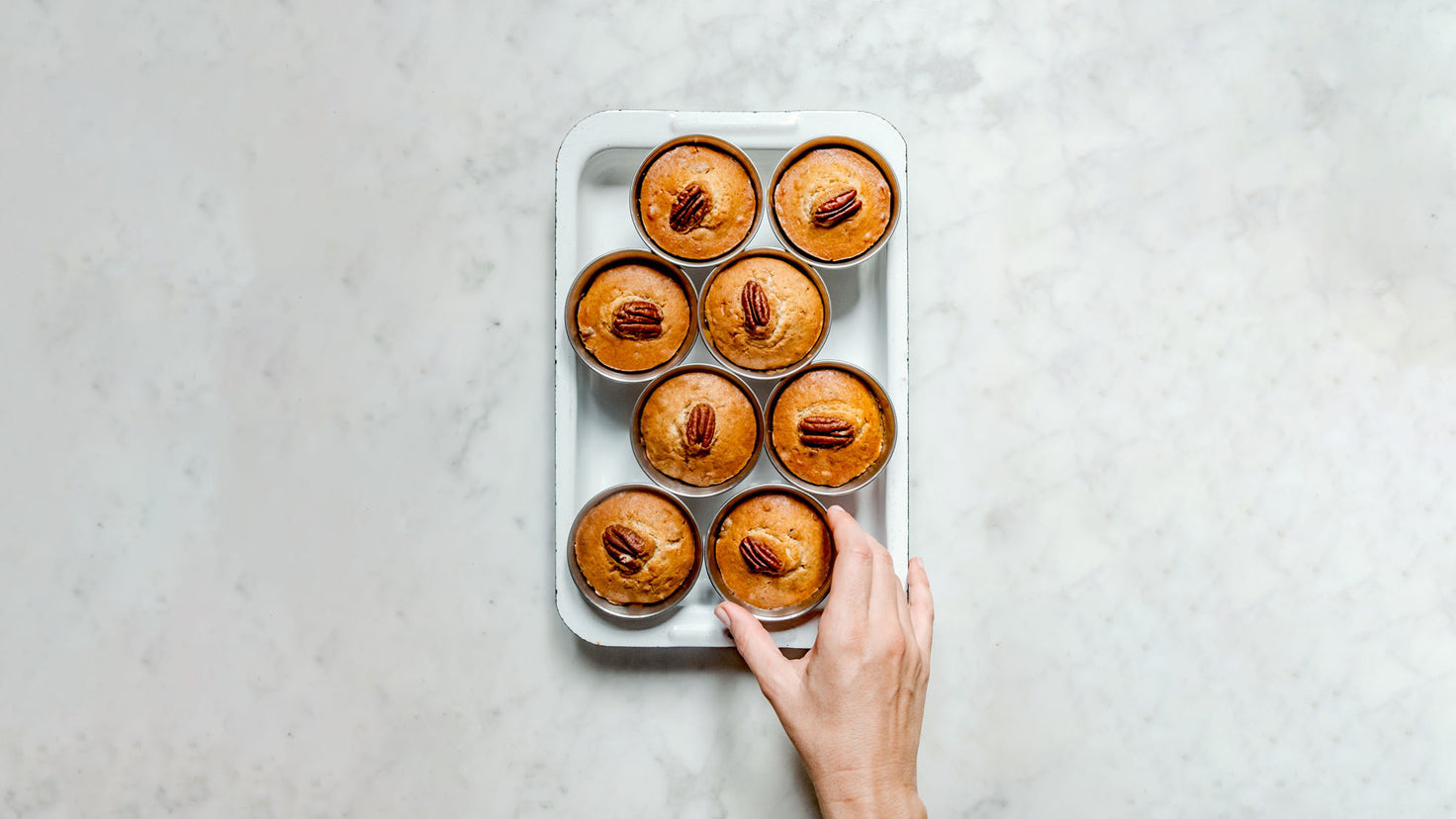 A hand reaches for a pecan-topped muffin, fresh from the oven in a muffin tin.