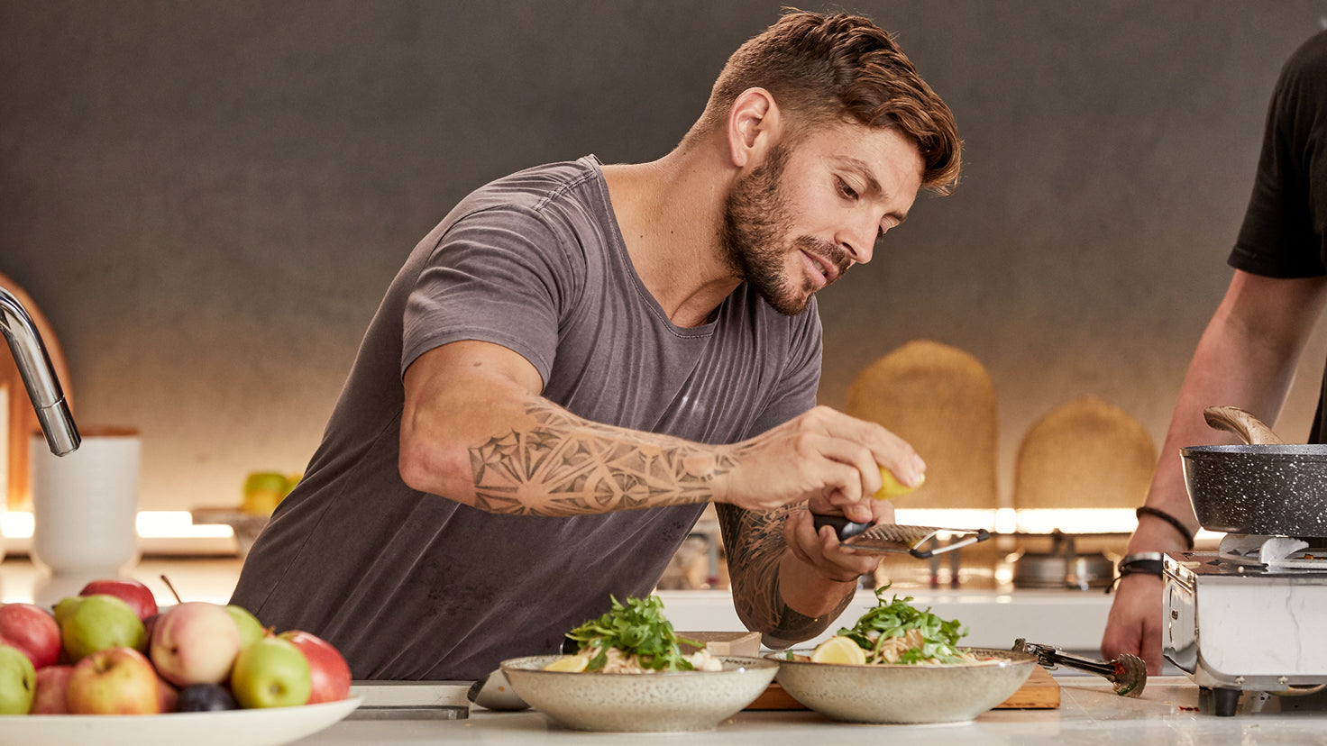 A man with tattoos grates lemon zest into a bowl of food.