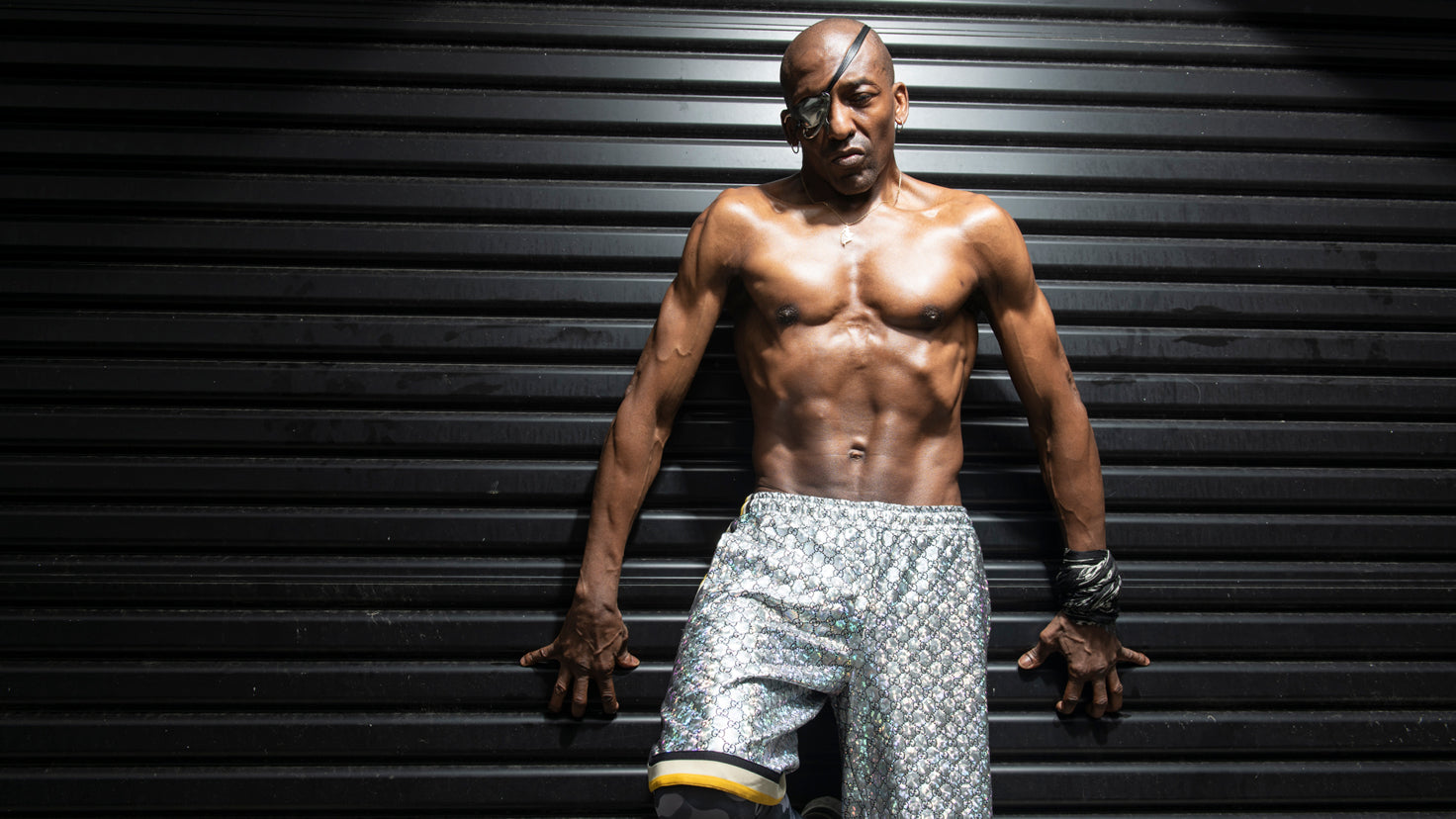 Michael Olajide Jr, shirtless with visible abs, leans against a corrugated roller door.