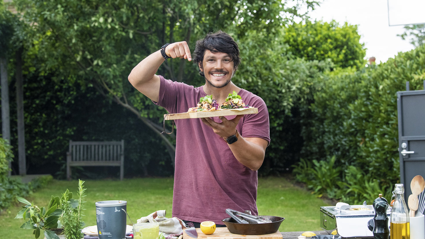 Dan Churchill holds a chopping board with two tacos while standing in a garden.