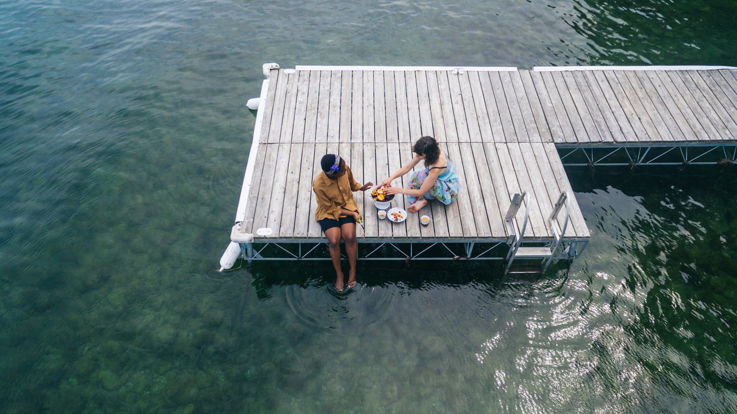 Two women share a picnic while sitting on a jetty over a lake.