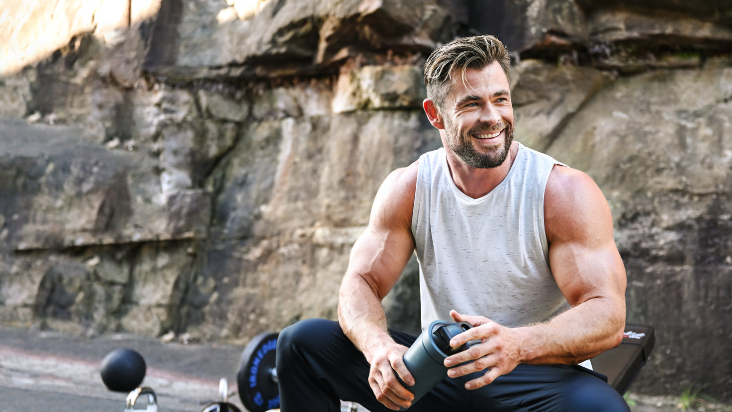 Chris Hemsworth rests between sets, sitting in front of a rocky wall and smiling off-camera