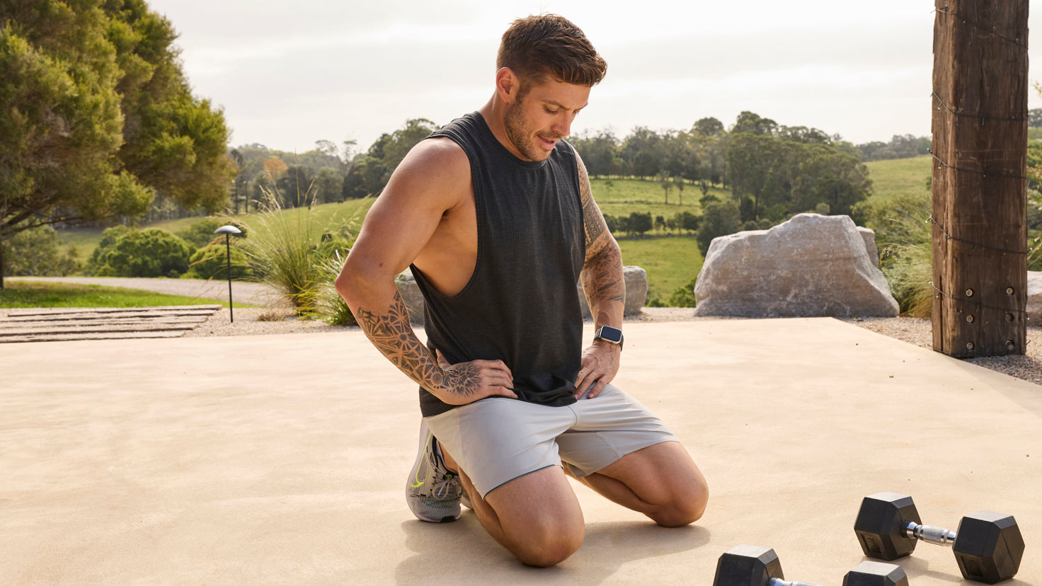 Centr trainer Luke Zocchi is kneeling on a wooden deck, hands on hips, looking down at a pair of dum