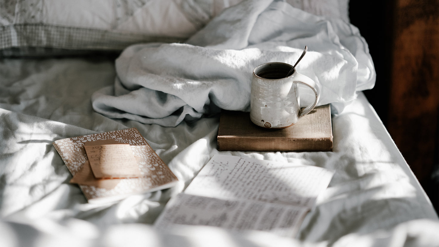 Books, notepads and a cup of coffee rest on an unmade bed with white linens.
