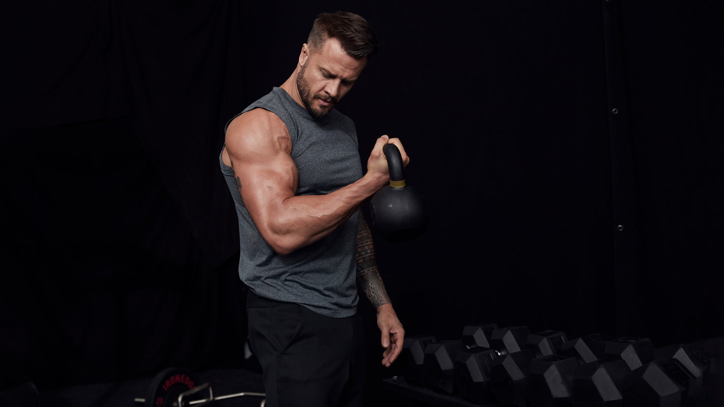 Centr trainer Bobby Holland Hanton performs a bicep curl against a black background in a gym.