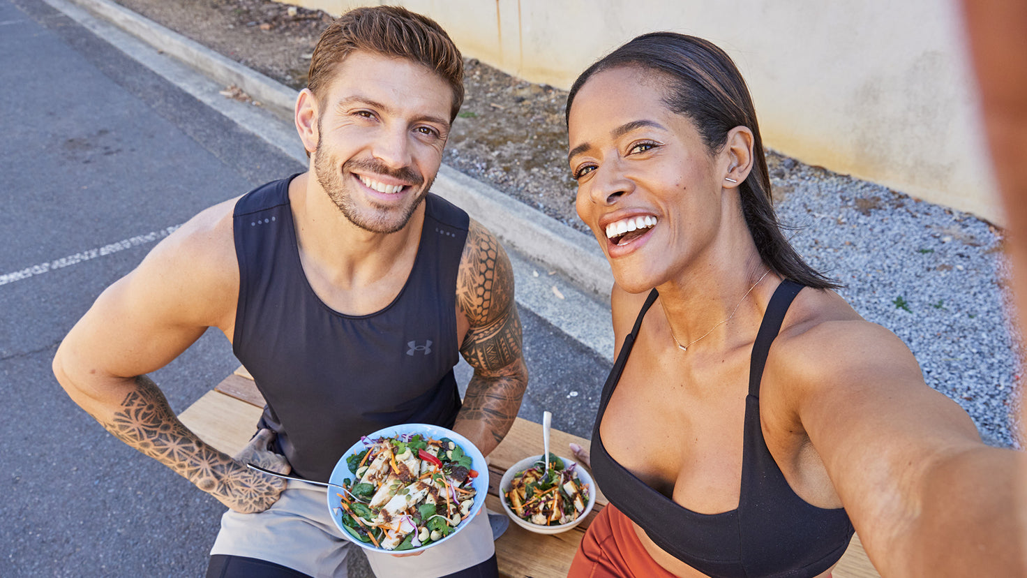 Centr trainers Ingrid Clay and Luke Zocchi pose for an overhead selfie, each holding a bowl of good