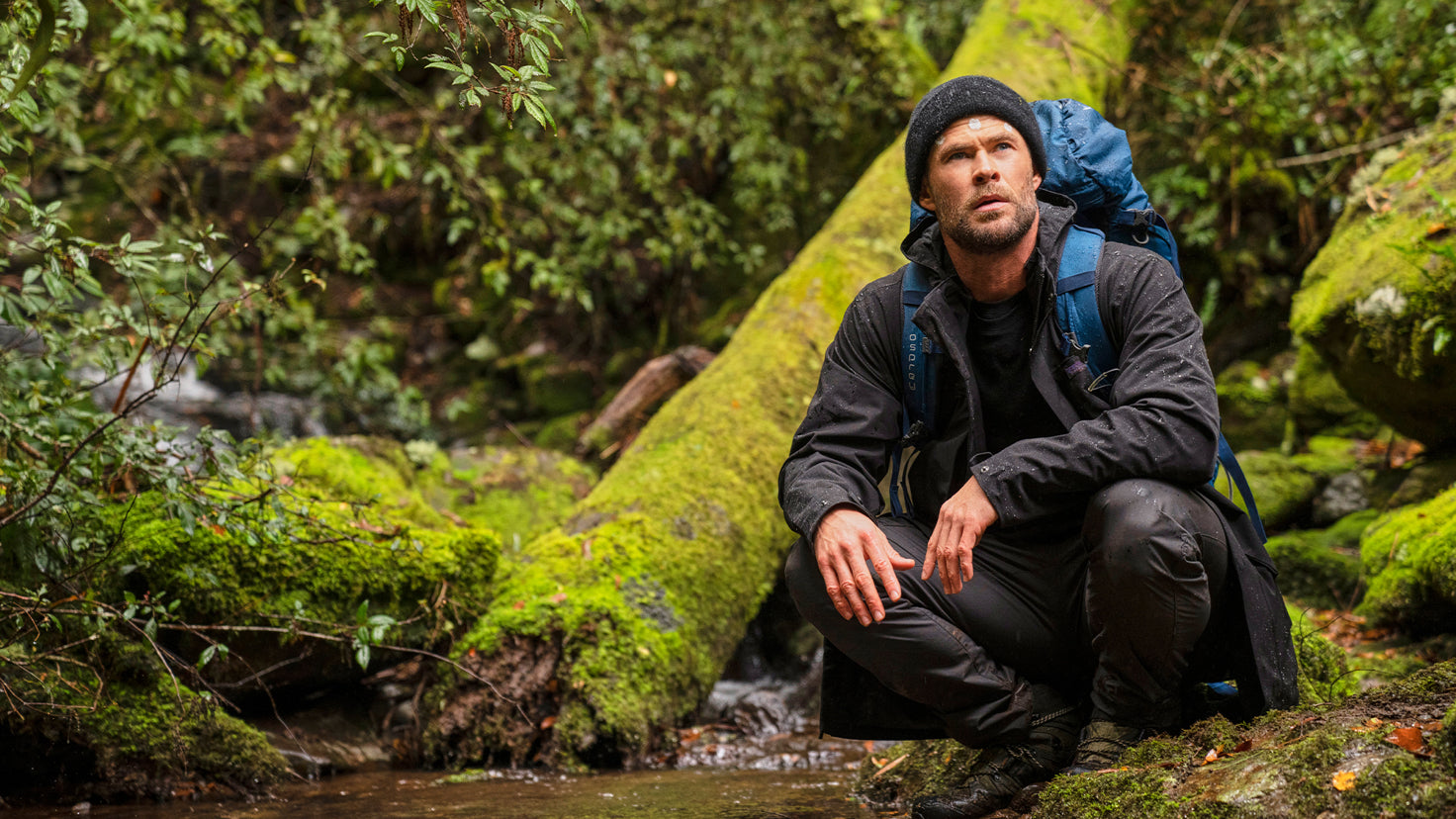 Centr founder Chris Hemsworth crouches on a mossy rock and looks off into the distance.