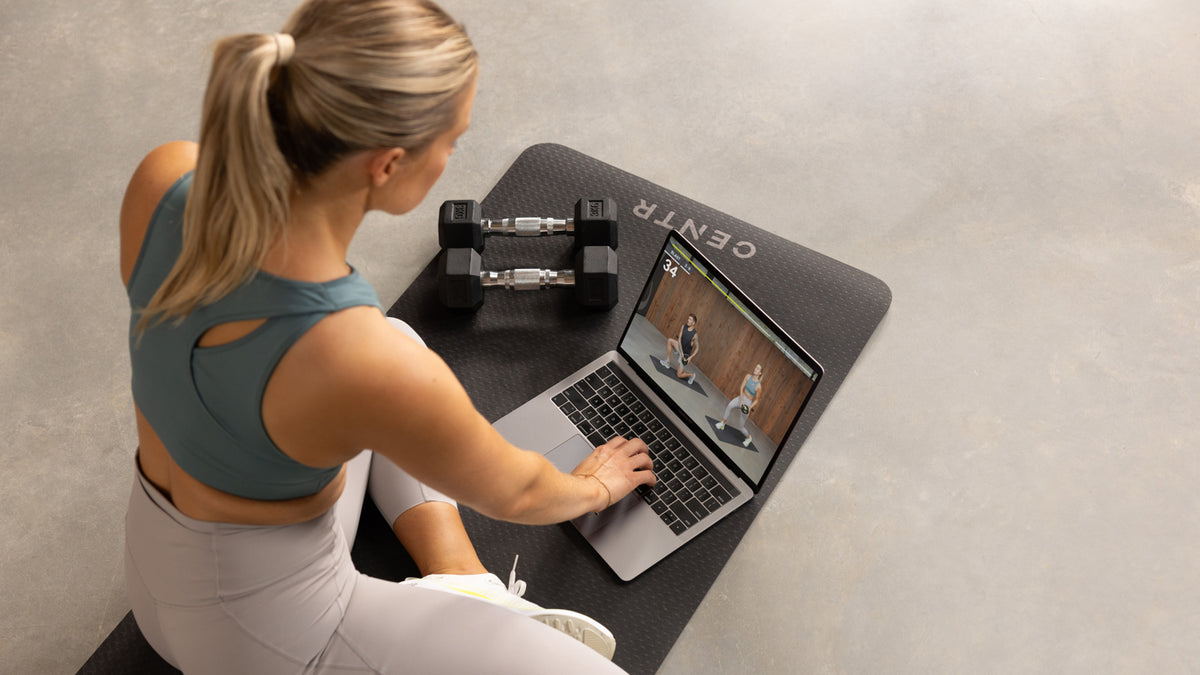 Centr trainer Alexz parvi sits on a yoga mat with dumbbells watching a Centr workout video on a lapt