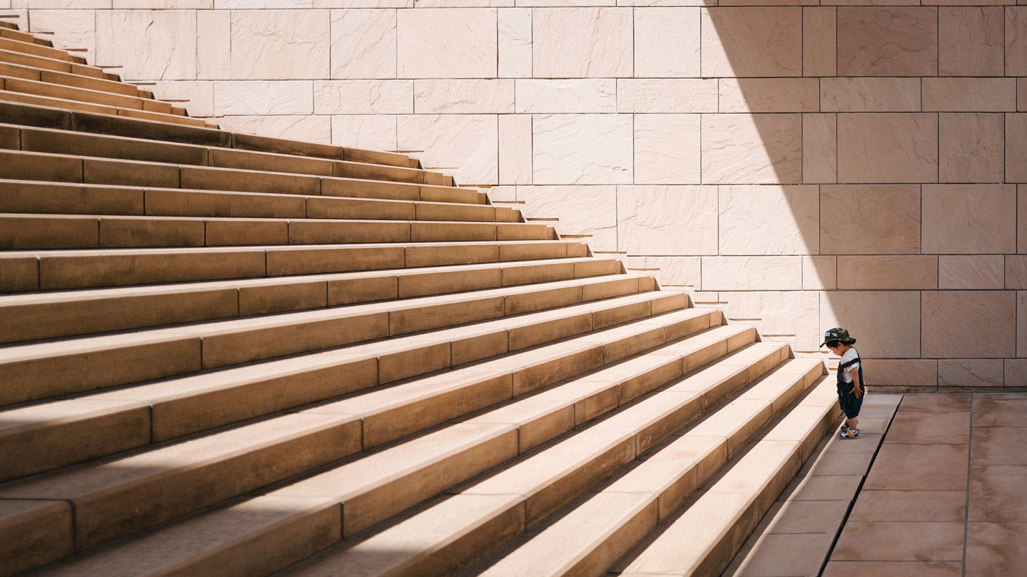 A toddler in overalls stands at the base of a large, intimidating set of stairs.