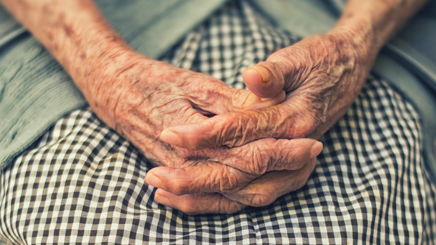 A woman's pair of aged hands, fingers interlaced, rest in in her lap. The edge of her green knitted