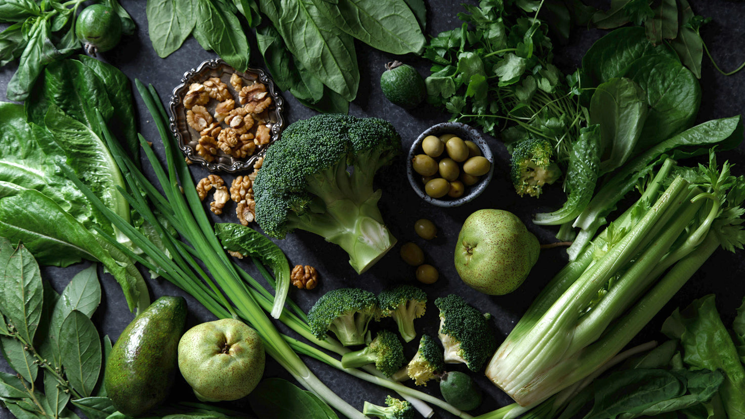 Green vegetables including broccoli, celery and spinach laid on a blue-grey bench.