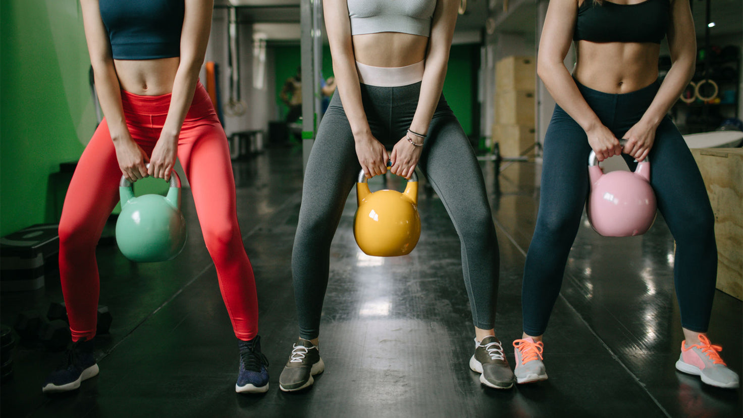 Three slim women lift colorful kettlebells in a gym to build muscle and strength.