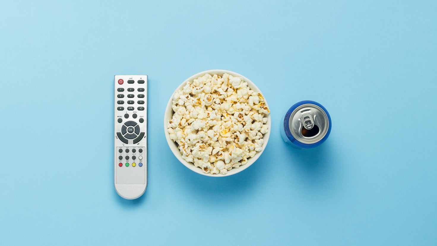 A TV remote, a bowl of popcorn and a can of soda.