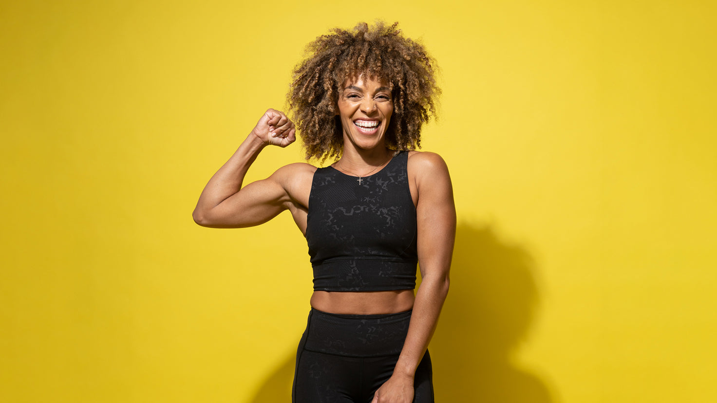 Centr HIIT Strength trainer Ashley Joi smiles and flexes her bicep against a yellow background.
