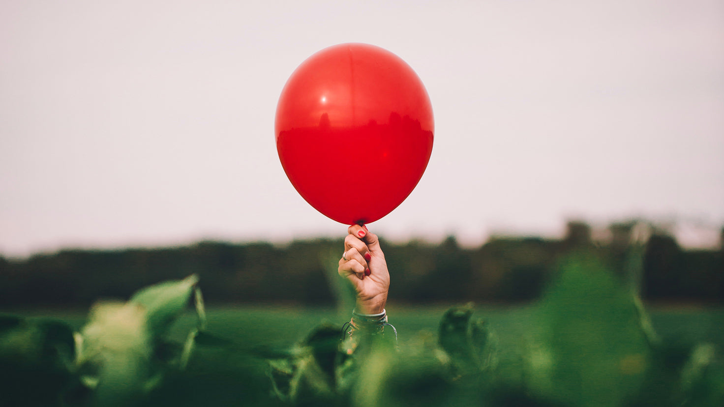A hand with red nail polish holds up a red balloon above a hedge.