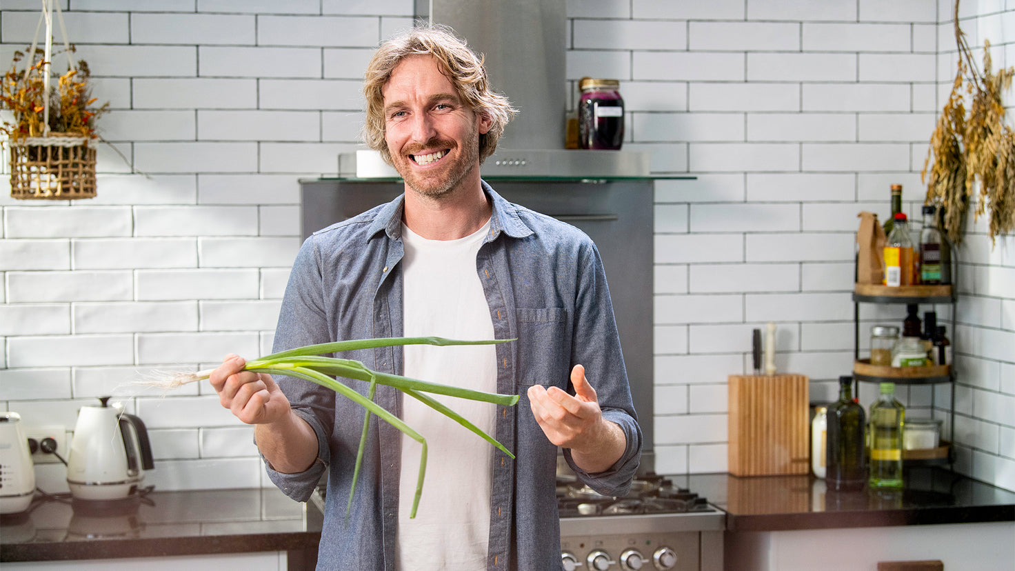 Darren Robertson, smiling in the kitchen, holds a raw whole spring onion in one hand.