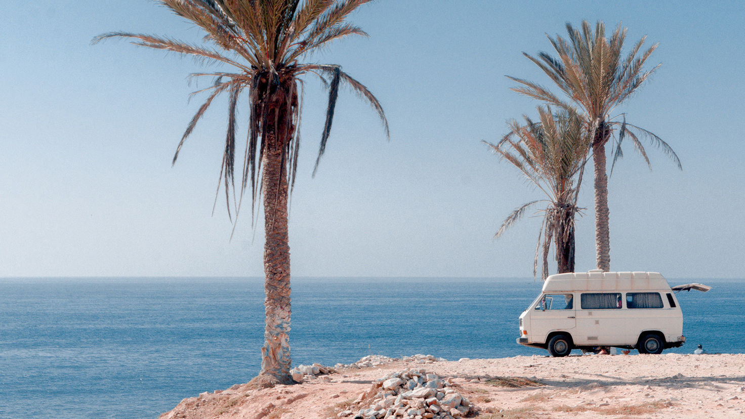 A white van parked on the edge of a rocky beach next to palm trees.