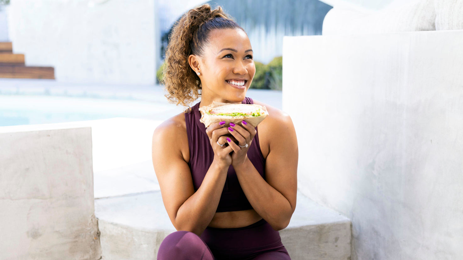 Centr trainer Maricris Lapaix, wearing gym wear, sits on a limestone step, holding half a salad sand