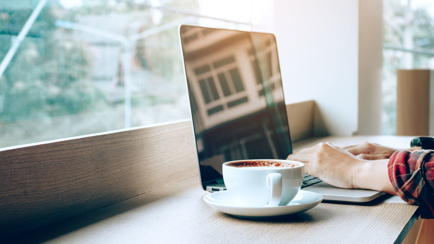 A close up of a person typing on a laptop with a coffee beside them in a cafe.