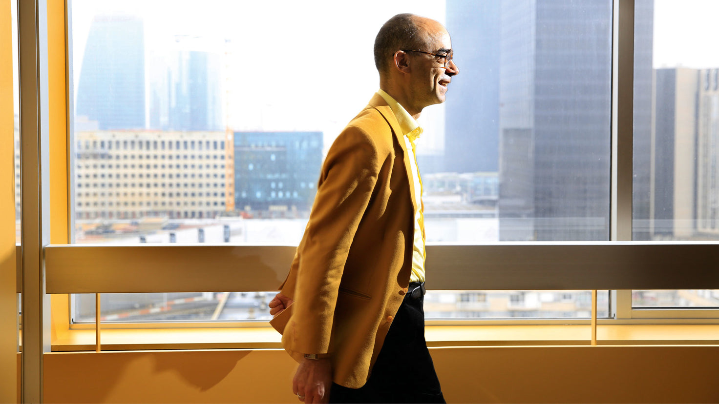 Fabrice Midal wearing a mustard-colored jacket, walking through a sunny hallway.