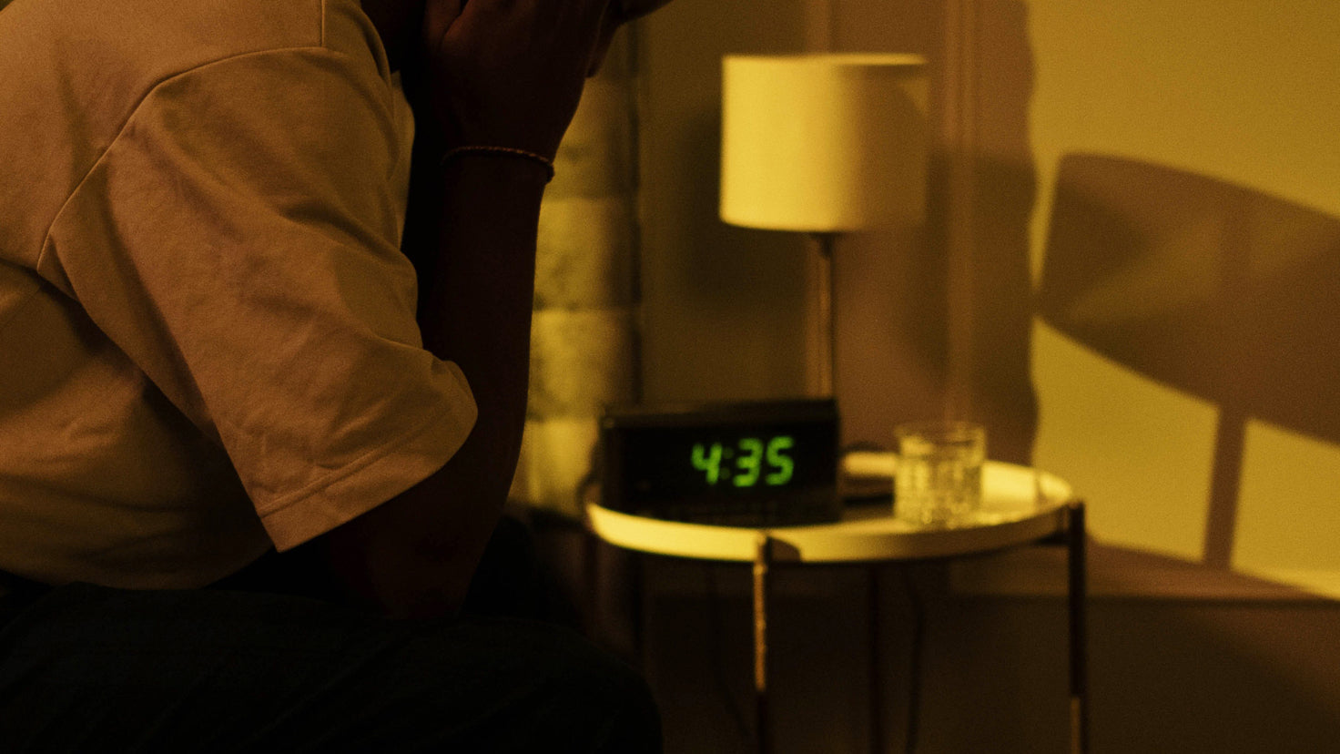 A person sits in a darkened room, head in hands. The digital clock in the bedside table in the backg