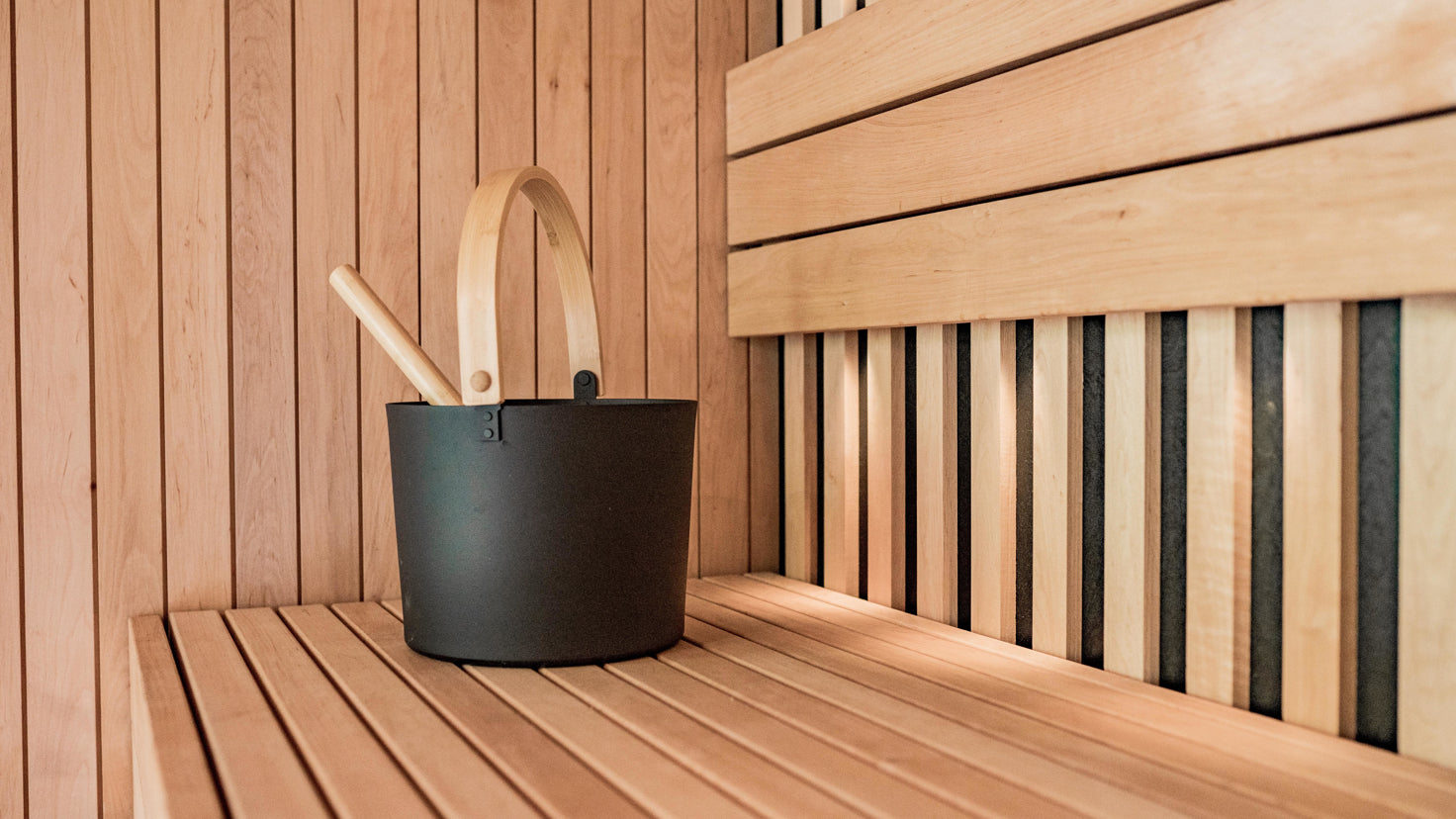 A black bucket with a pine handle sits on a pine bench inside a pine-clad sauna.