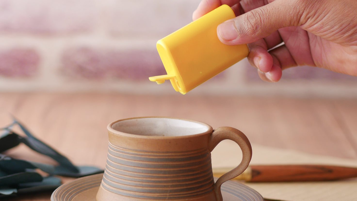 An open sweetener dispenser is held over a stripey brown pottery mug.