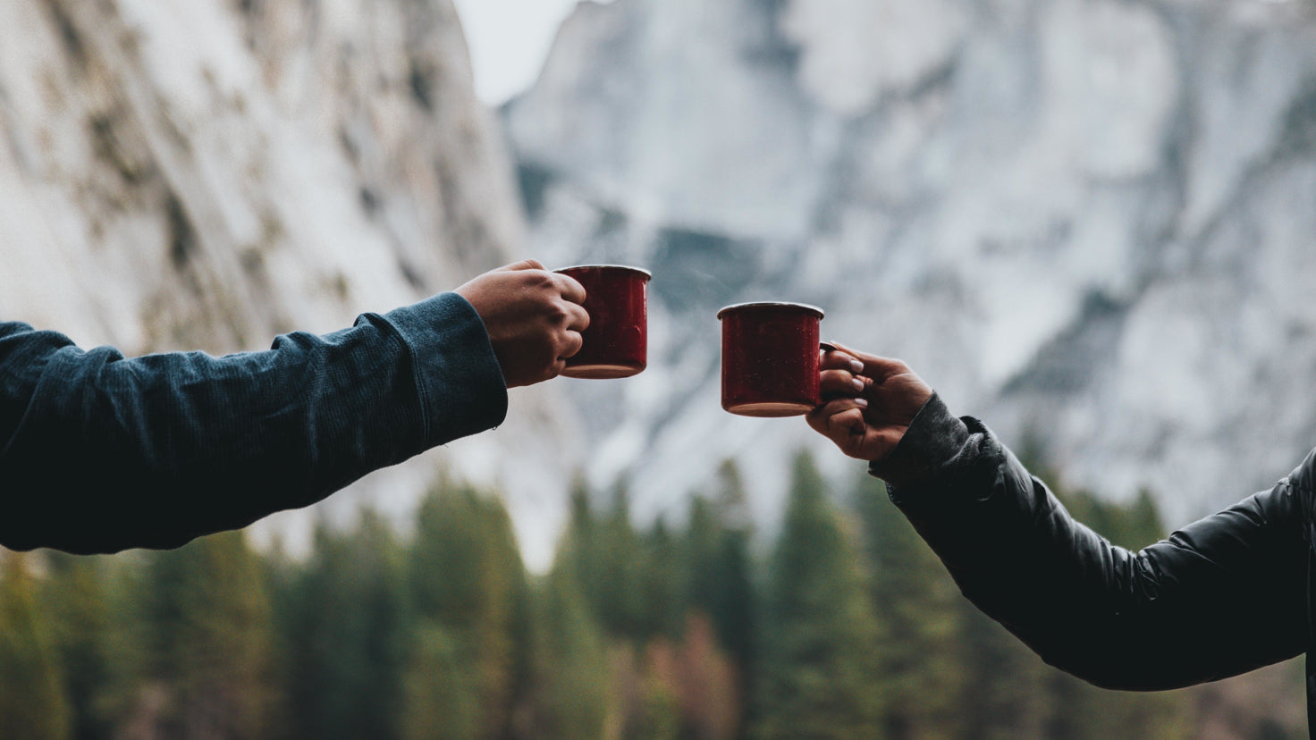 Two arms, with mugs in hand, reach out to each other, with a mountain view in the background.