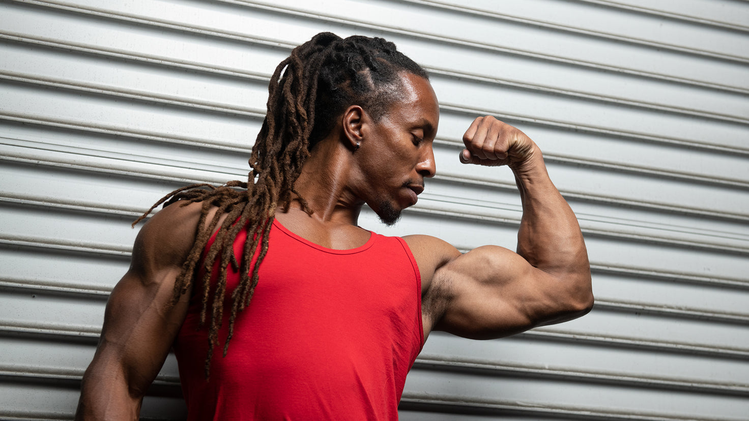 A muscular bodybuilder with dreadlocks flexes his bicep while looking at it admiringly.