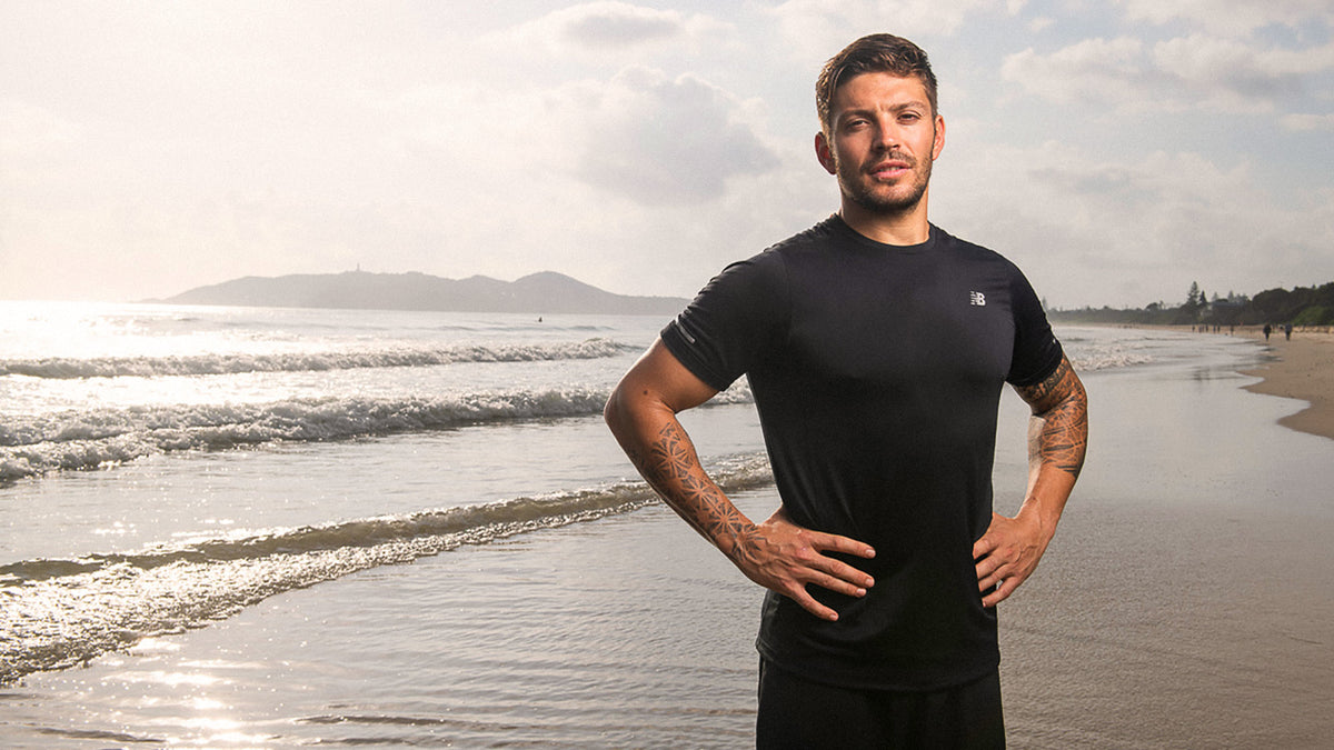 A man with tattoos (Luke Zocchi) stands on a beach.