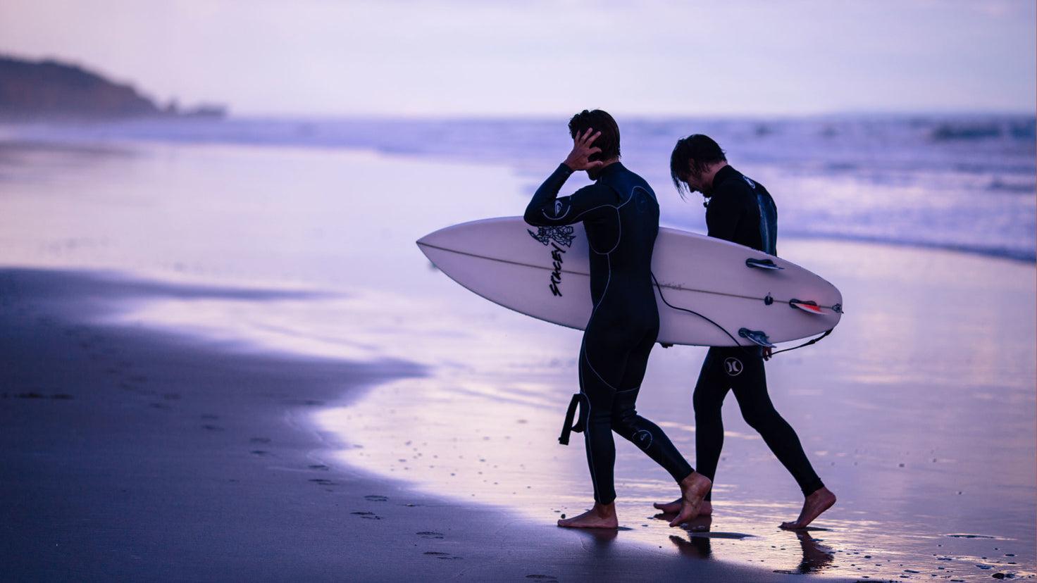 Two men wearing wetsuits, one holding a surfboard, walk on the beach at dawn.