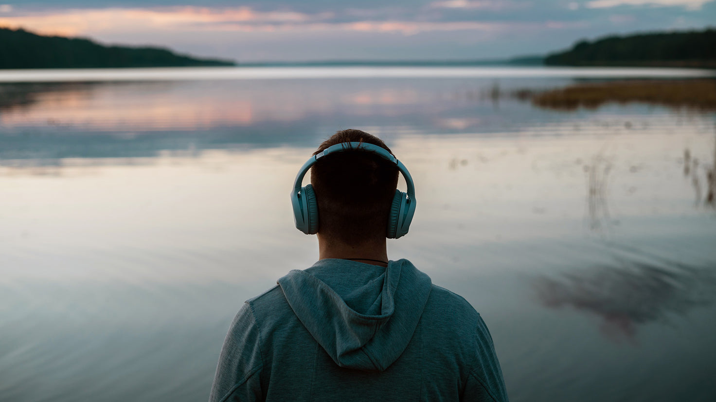 A man wearing headphones looks out over a picturesque lake.